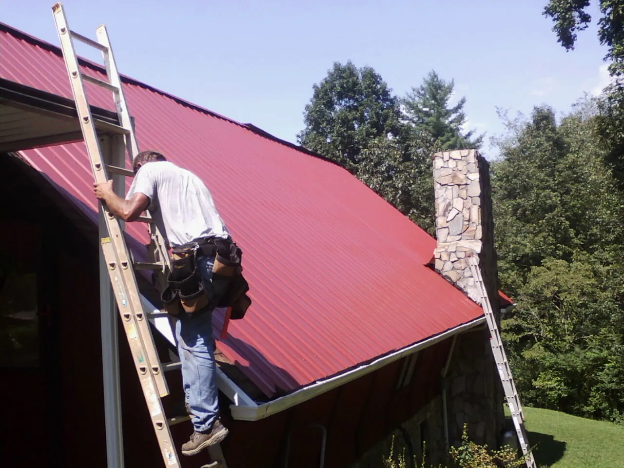 Roofer on a ladder beside a red metal roof and stone chimney — Soffit & Fascia Repair by Roofing Brothers in Princess Anne